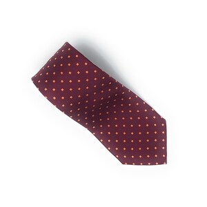 May include: A burgundy tie with a repeating pattern of small, orange squares and blue dots. The tie is made of a smooth fabric and is angled across a white background. The tie is a classic accessory for formal or business attire.