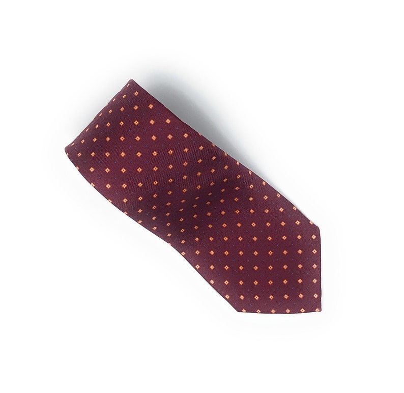 May include: A burgundy tie with a repeating pattern of small, orange squares and blue dots. The tie is made of a smooth fabric and is angled across a white background. The tie is a classic accessory for formal or business attire.
