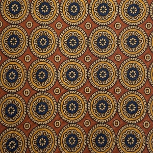 May include: Close-up of a patterned fabric featuring repeating circular designs. Each circle has a dark blue center, surrounded by yellow and cream-colored floral patterns. The background is a warm brown color.