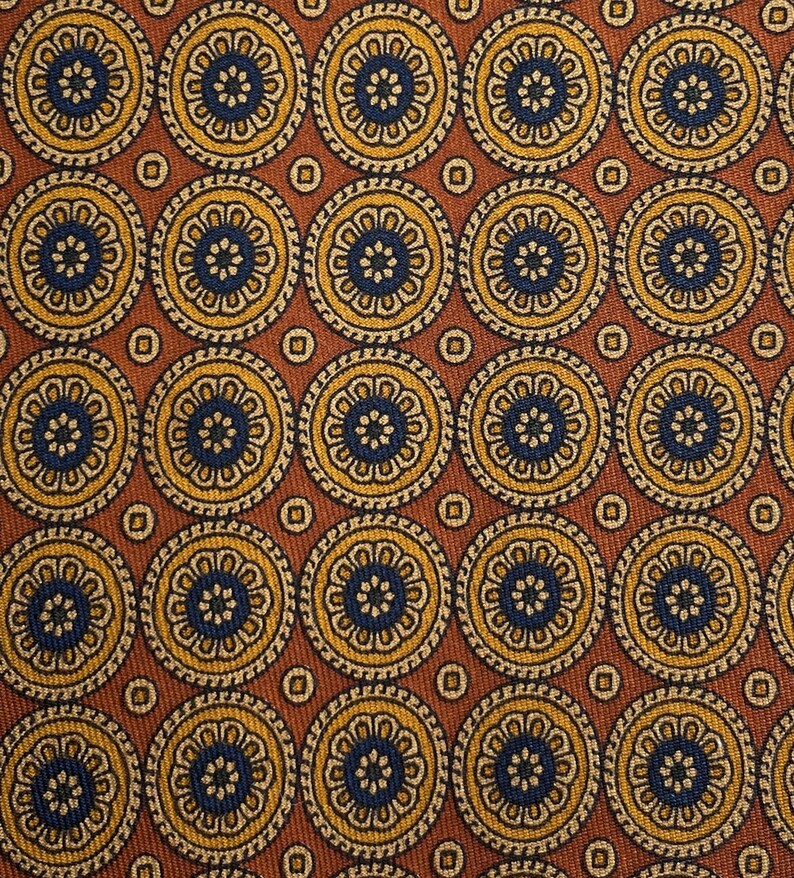 May include: Close-up of a patterned fabric featuring repeating circular designs. Each circle has a dark blue center, surrounded by yellow and cream-colored floral patterns. The background is a warm brown color.