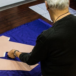 May include: A person is cutting out fabric pieces on a blue table cloth. The fabric pieces are beige and the person is wearing a black shirt.