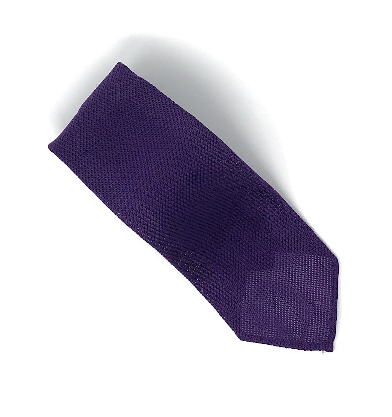 May include: A solid purple necktie with a textured weave. The tie is displayed diagonally against a white background. The tie's color is a deep, rich shade of purple.