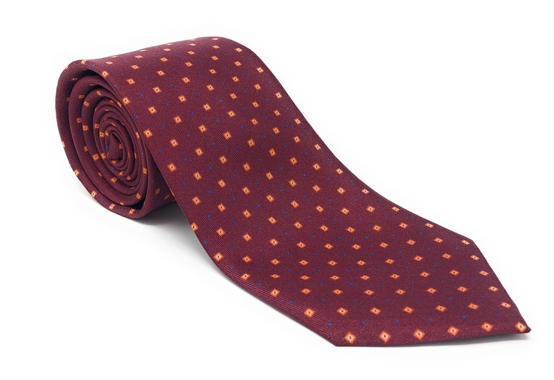 May include: A rolled burgundy tie with a repeating pattern of small orange squares and blue dots. The tie is made of a textured fabric and is displayed on a white background. This classic tie is suitable for formal or business attire.