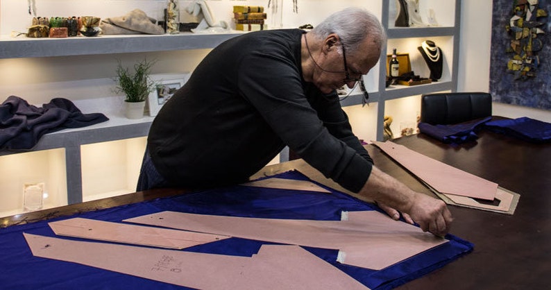 May include: A person is working on a blue fabric with brown paper patterns. The person is wearing a black long-sleeved shirt and glasses. The fabric is laid out on a wooden table.