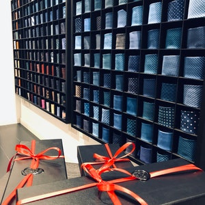 May include: A stack of black gift boxes with red ribbon bows. The boxes are arranged in a pyramid shape. The boxes are in front of a wall with a display of blue and white patterned ties.