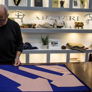 May include: A man in a black shirt is working on a blue fabric with brown paper pattern pieces. The fabric is laid out on a wooden table. The man is standing in front of a shelf with various items, including a sign that says "ARCIERI".