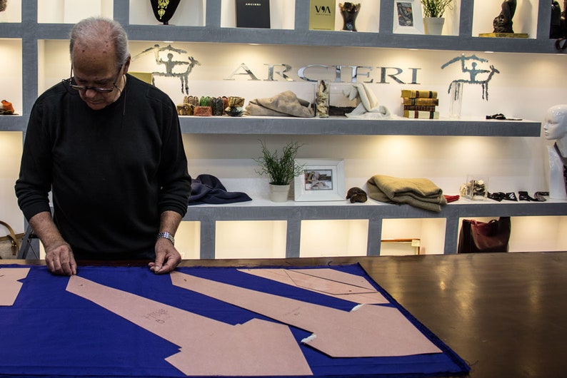 May include: A man in a black shirt is working on a blue fabric with brown paper pattern pieces. The fabric is laid out on a wooden table. The man is standing in front of a shelf with various items, including a sign that says "ARCIERI".