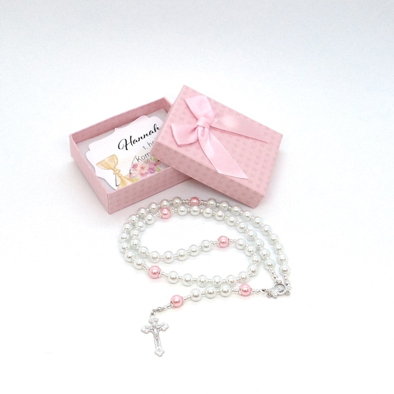 May include: A white pearl rosary with pink accent beads and a silver crucifix lies next to a pink gift box tied with a matching bow. A card inside the box reads "Hannah 1. he Komunia".