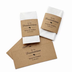 Puede incluir: Pañuelos de papel blancos con bandas de papel marrón. Las bandas están impresas con el texto "für die Freudentränen", los nombres "Sonja & Matteo" y la fecha "22.09.2017". Se ven varias bandas y pañuelos.