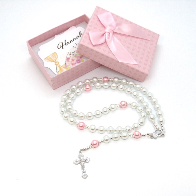 May include: A rosary with white and pink beads, and a silver crucifix. The rosary is displayed with a pink gift box tied with a satin bow. A card inside the box has text and a floral design.
