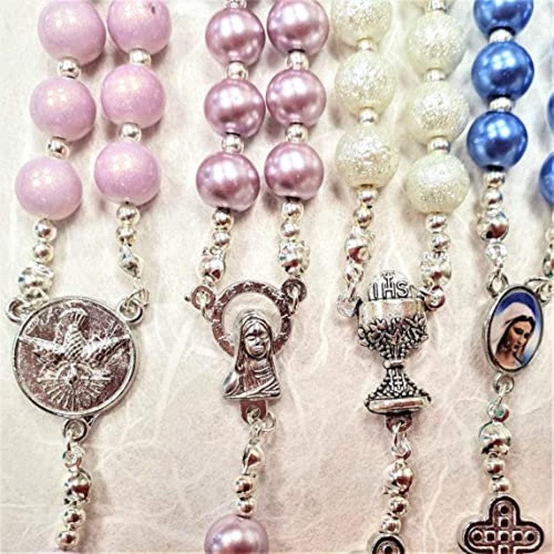 May include: Close-up of several rosaries with different colored beads, including pink, purple, white, and blue. Each rosary has a silver-toned metal pendant with religious imagery, such as a dove, a chalice, and a depiction of a saint.
