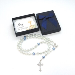 May include: A pearl rosary with blue beads and a silver crucifix lies next to a black gift box with a blue bow. A card inside the box reads "Leny zur Taufe am 17 Juli 2021". The rosary is a religious item, often used for prayer.