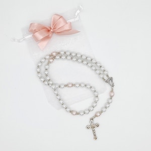 Puede incluir: Un rosario de perlas blancas con una cruz de plata y una bolsa de sat&eacute;n rosa. El rosario est&aacute; hecho de perlas blancas y cuentas de plata.