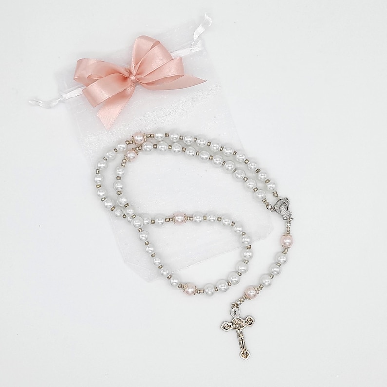 Puede incluir: Un rosario de perlas blancas con una cruz de plata y una bolsa de sat&eacute;n rosa. El rosario est&aacute; hecho de perlas blancas y cuentas de plata.