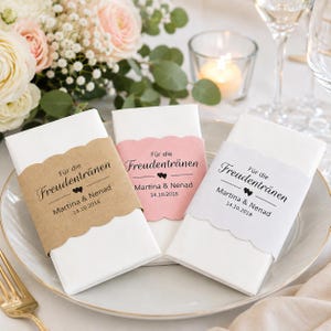 May include: Three white folded napkins with decorative paper bands. The bands are in brown, pink, and white, each with the text "Für die Freudentränen" and names and a date. The napkins are on a white plate, with flowers and a candle in the background.