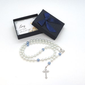 May include: A pearl rosary with blue accents and a silver crucifix, presented in a black gift box with a blue ribbon. The box contains a card with the name "Leny" and the text "zur Taufe am 17 Juli 2021".