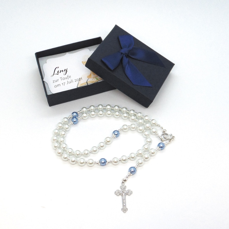 May include: A pearl rosary with blue accents and a silver crucifix, presented in a black gift box with a blue ribbon. The box contains a card with the name "Leny" and the text "zur Taufe am 17 Juli 2021".