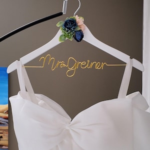 Könnte beinhalten: Ein weißer Holzkleiderbügel mit einem goldenen Drahtschriftzug Mrs. Greiner, verziert mit einem Bündel aus künstlichen dunkelblauen und hellrosa Blumen mit grünen Blättern, an dem ein weißes Kleid mit einem großen schleifenartigen Oberteil hängt.