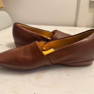 May include: A pair of brown leather slip-on shoes with a pointed toe. The shoes have a low heel and a yellow interior. The leather appears to be smooth and has a slight sheen. The shoes are likely for casual wear.