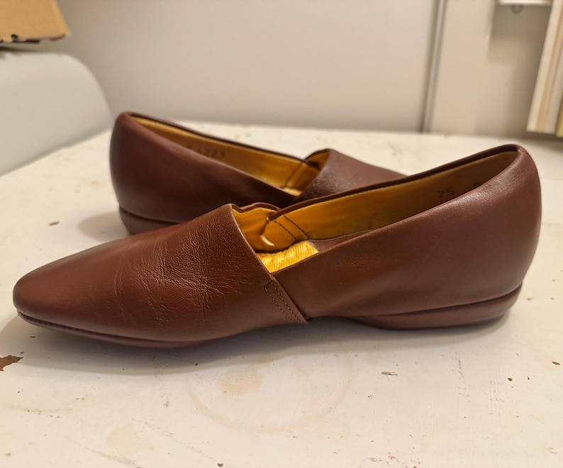 May include: A pair of brown leather slip-on shoes with a pointed toe. The shoes have a low heel and a yellow interior. The leather appears to be smooth and has a slight sheen. The shoes are likely for casual wear.
