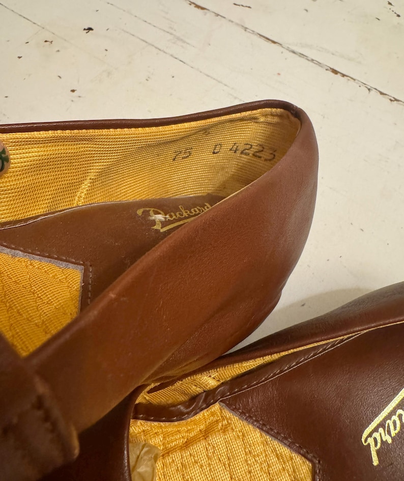 May include: Close-up of a pair of brown leather shoes. The interior lining is yellow. The shoes have the brand name "Packard" printed on the insole. The size is marked as "7.5 D 4223".