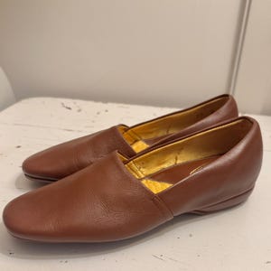 May include: A pair of brown leather loafers. The shoes have a rounded toe and a low heel. The interior lining is a golden yellow color. The shoes are sitting on a white surface.
