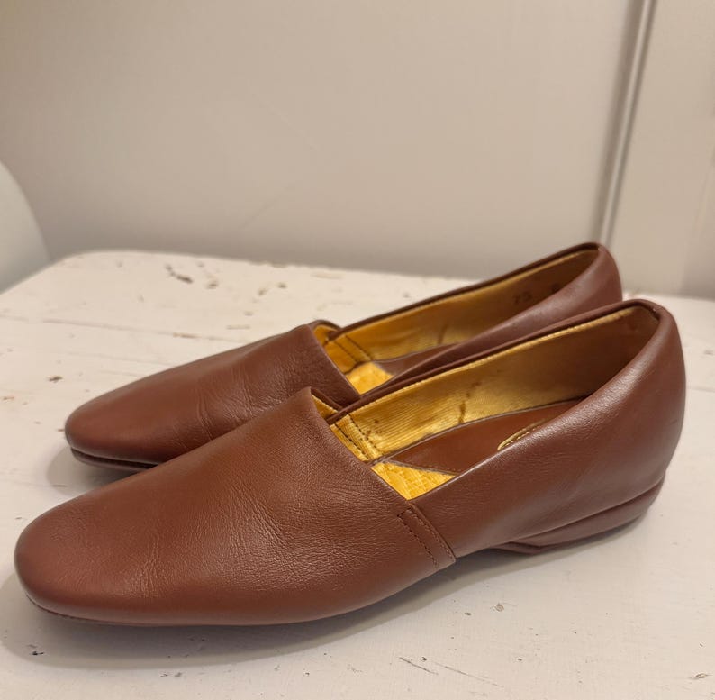 May include: A pair of brown leather loafers. The shoes have a rounded toe and a low heel. The interior lining is a golden yellow color. The shoes are sitting on a white surface.