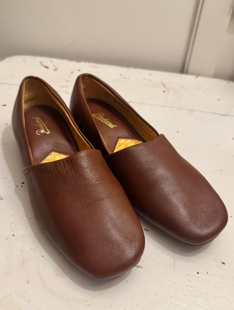 May include: A pair of brown leather slip-on shoes. The shoes have a rounded toe and a low heel. The interior is lined with a yellow material. The brand name "Packard" is visible inside the shoe.