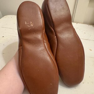 May include: A pair of brown leather slippers. The soles are visible, showing the shape and stitching. The leather has a smooth texture and a warm, reddish-brown color. The slippers appear to be well-worn.