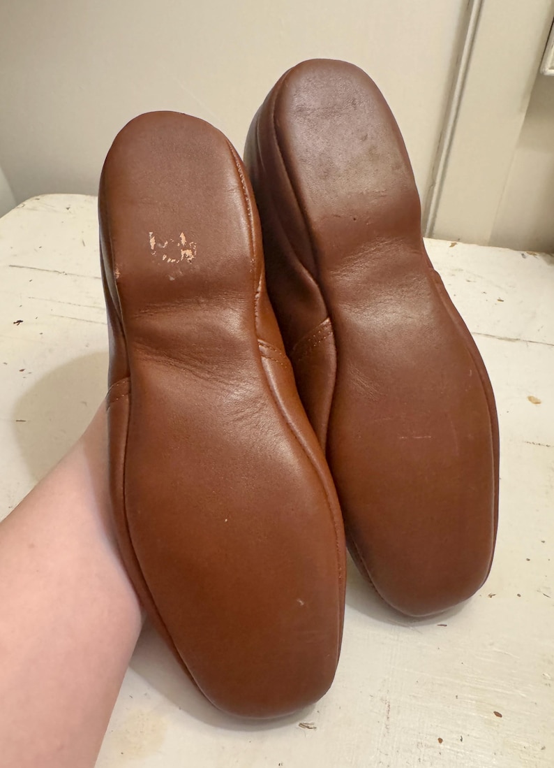 May include: A pair of brown leather slippers. The soles are visible, showing the shape and stitching. The leather has a smooth texture and a warm, reddish-brown color. The slippers appear to be well-worn.