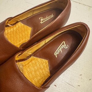 May include: A pair of brown leather loafers with the brand name "Packard" embossed in gold lettering inside. The interior lining is a textured yellow fabric. The shoes are in a close-up shot, showcasing the details of the leather and stitching.
