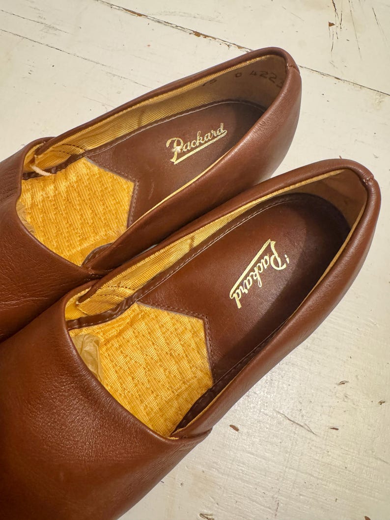 May include: A pair of brown leather loafers with the brand name "Packard" embossed in gold lettering inside. The interior lining is a textured yellow fabric. The shoes are in a close-up shot, showcasing the details of the leather and stitching.