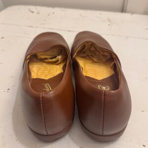May include: A pair of brown leather slip-on shoes. The shoes have a rounded toe and a low heel. The interior is lined with a gold-colored material. The shoes are likely for indoor use, such as slippers or house shoes.