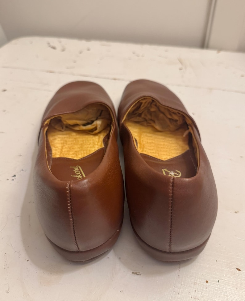 May include: A pair of brown leather slip-on shoes. The shoes have a rounded toe and a low heel. The interior is lined with a gold-colored material. The shoes are likely for indoor use, such as slippers or house shoes.