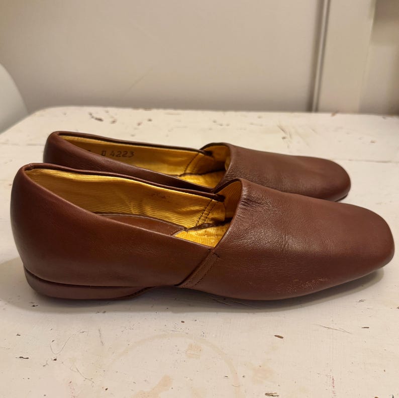 May include: A pair of brown leather loafers with a rounded toe. The interior lining is gold. The shoes are on a white surface. The shoes appear to be unworn.