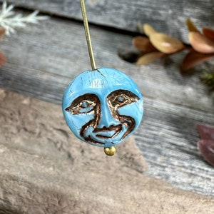 May include: A handmade bead with a celestial design. The round, light blue bead features a detailed face with brown accents and gold-colored eyes. It is attached to a long, thin, gold-colored pin, perfect for jewelry making or crafting.