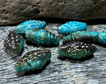 May include: A collection of small, decorative beads with a textured, ridged surface. The beads are primarily turquoise and brown, with a metallic sheen. They are arranged on a weathered, gray wooden surface.