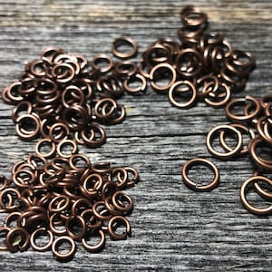 May include: Four piles of copper jump rings on a wooden surface. The jump rings are small and round, and they are arranged in a semi-circle.