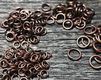 May include: Four piles of copper jump rings on a wooden surface. The jump rings are small and round, and they are arranged in a semi-circle.