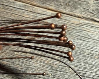 May include: Close-up of a bundle of copper-colored head pins. Each pin has a small, round bead at the top. The pins are arranged against a weathered, wooden surface, creating a rustic aesthetic. These pins are ideal for jewelry making and craft projects.