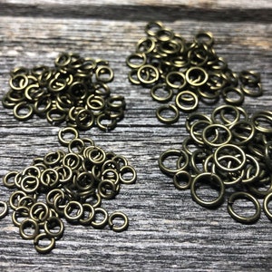 May include: Three piles of small, gold-colored jump rings on a wooden surface. The jump rings are about 5mm in diameter.
