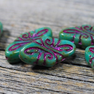 May include: Close-up of several teal-colored oval beads with a raised, magenta-colored swirl design. The beads have a glossy finish and are arranged on a weathered wooden surface. These beads are ideal for jewelry making.