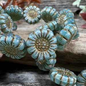 May include: A strand of turquoise blue flower-shaped beads with a distressed finish and gold-toned detailing in the center and along the petals. The beads are arranged in a circular pattern, showcasing a textured surface and a consistent floral design.
