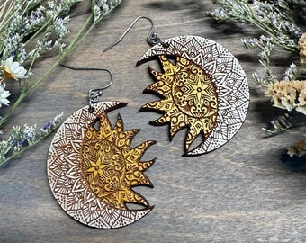 May include: A pair of circular wooden earrings featuring a laser-engraved sun and moon design. The sun is depicted in a golden yellow hue with intricate mandala patterns, while the surrounding crescent moon is white with detailed filigree.