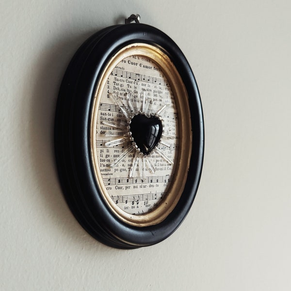 Oval black frame with black obsidian votive heart and quartz. Exvoto decoration. Old music sheet.