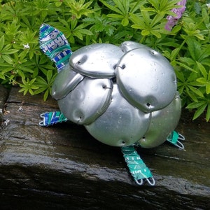 May include: Two small turtle sculptures made from aluminum can tabs. The turtles are silver with green and blue accents. They are sitting on a wooden surface.