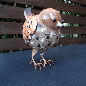 Op de afbeelding: Een metalen vogelbeeldje, geschilderd in bruin, wit en zwart. De vogel heeft een lange snavel en kleine, ronde ogen.