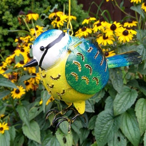 Op de afbeelding: Een blauwe vogel tuin decoratie gemaakt van metaal met een geel lichaam, blauwe vleugels en een wit hoofd. De vogel heeft een uitgesneden ontwerp en hangt aan een touwtje.