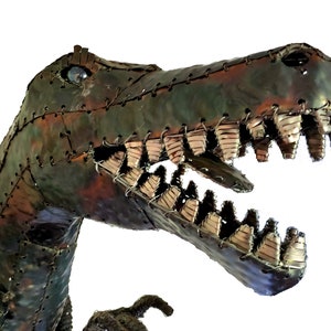 May include: Close-up of a metal dinosaur sculpture with a textured, green, brown, and silver finish. The dinosaur's mouth is open, revealing rows of teeth made of metal.