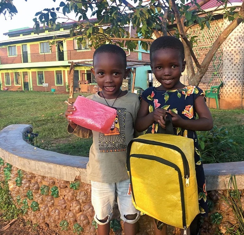 Pu&ograve; includere: Due bambini, uno con una borsa rosa e l'altro con uno zaino giallo, stanno in piedi davanti a un edificio fatto di bottiglie di plastica riciclate. Il bambino che porta lo zaino indossa un vestito blu con fiori. Il bambino che porta la borsa indossa una maglietta grigia con la scritta "King of the Jungle" stampata.